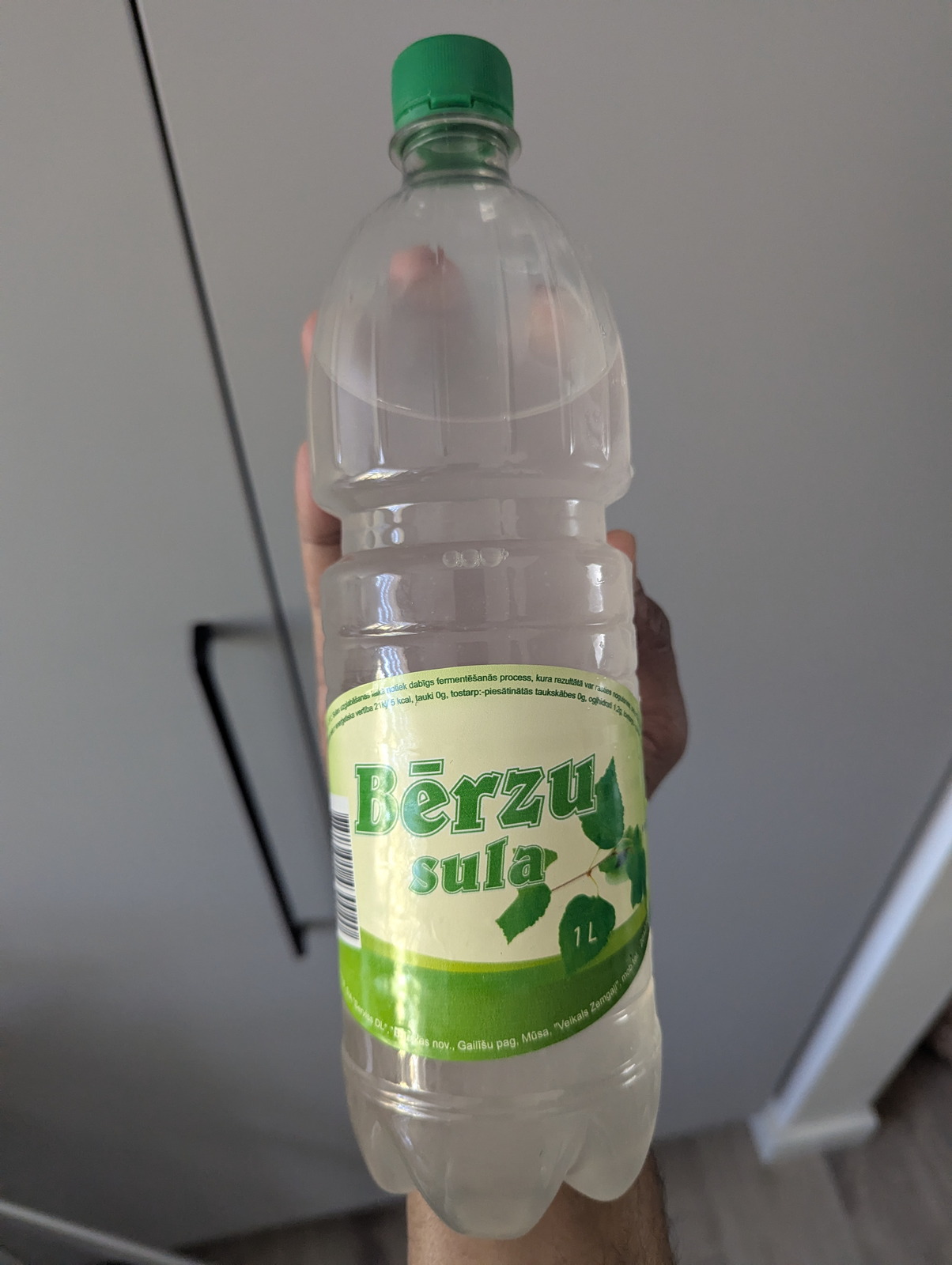 Una botella PET de 1 litro de Bērzu sula (savia de abedul letona) con etiqueta de hojas verdes, sostenida contra una pared de cocina